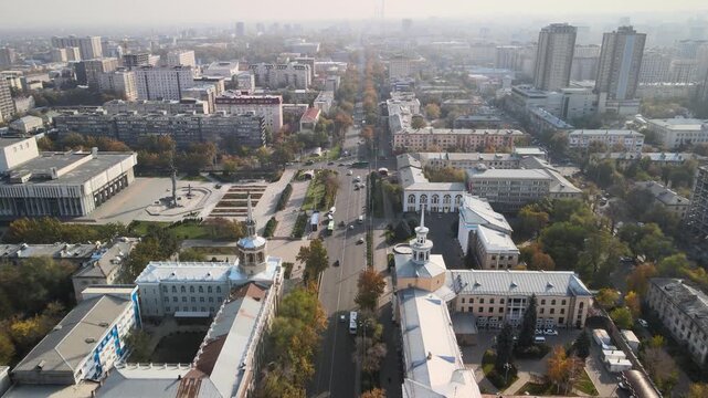 Drone flyover of Soviet-era institutional architecture along Chuy Avenue in Bishkek, Kyrgyzstan. Symmetrical buildings with rotunda elements surrounded by autumn trees and city traffic.