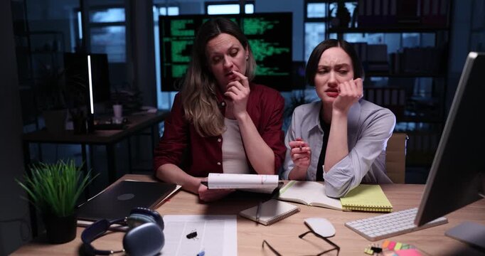 Businesswomen study documents and prepare report on computer late at night. Females compare business documents with digital information on computer