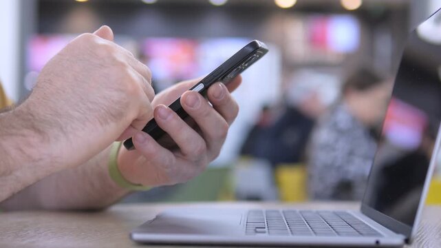 Hand types on smartphone beside open laptop. Focus on fingers scrolling or tapping screen. Background blurred with cafe or office setting. Person appears focused, multitasking digitally