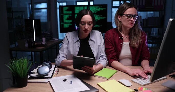 Women office workers compare information on computer and tablet. Tired female managers prepare detailed reports late at night in empty office