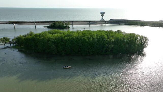 Drone video capturing a small motorboat sailing on the river estuary next to a green island and pedestrian bridge in the coastal resort of Anaklia, Georgia, with the sun reflecting on the water.