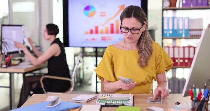 Focused woman accountant counts dollar bills using calculating machine at table. Blonde office worker calculates exact amount money in modern office