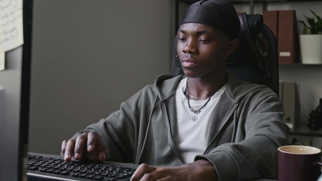 Tilt up shot of young African American programmer typing on keyboard, holding baseball while thinking in front of computer screen. Clip for tech startups, software development, and remote work ads