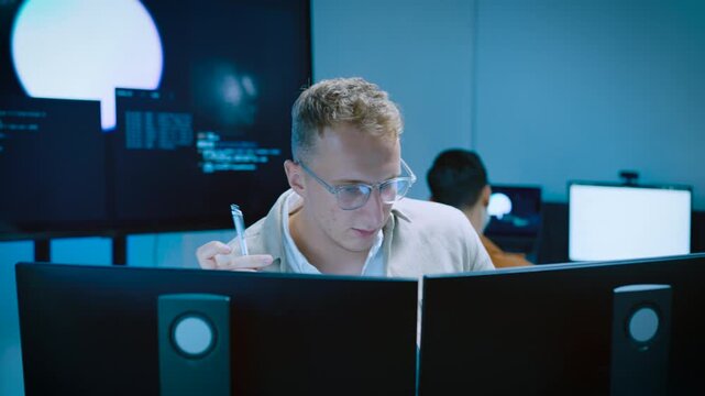 Pensive young male programmer holding pen while thinking of programming code idea and analyzing data on computer in tech office