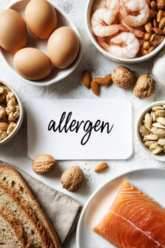A variety of potential food allergens are arranged on a white background. Eggs, salmon, shrimp, peanuts, and nuts surround a card with the word allergen