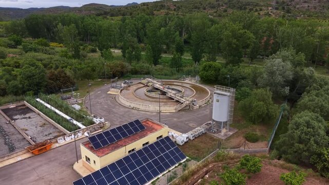 Compact wastewater treatment plant surrounded by green trees, featuring circular clarifier tank, solar panels on facility roof, sedimentation basins and storage silo in rural landscape