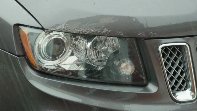 Car headlight covered with rain water during heavy downpour. Wet vehicle front end with water droplets on headlamp and hood in stormy weather. Automotive detail under intense rainfall with reduced