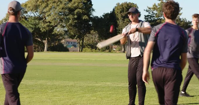 Diverse friends bowling ball to batsman tapping cricket bat, practicing batting in park