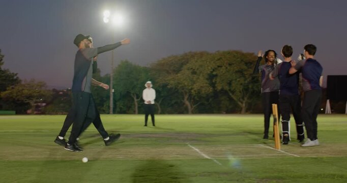 Male fielders spotting ball sprinting to uproot bails, celebrating wicket on floodlit pitch