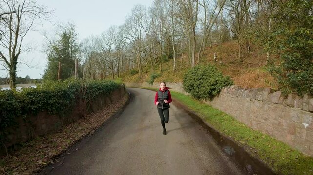 Cardio inspired middle aged woman running along curved countryside road beside stone wall and woodland landscape. Activating metabolism, improving circulation. Building resilience, enhancing mood
