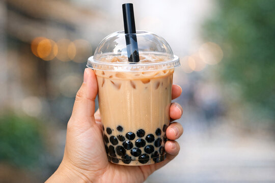 A person's hand is shown holding a cup of iced tea with tapioca balls on the street at night.