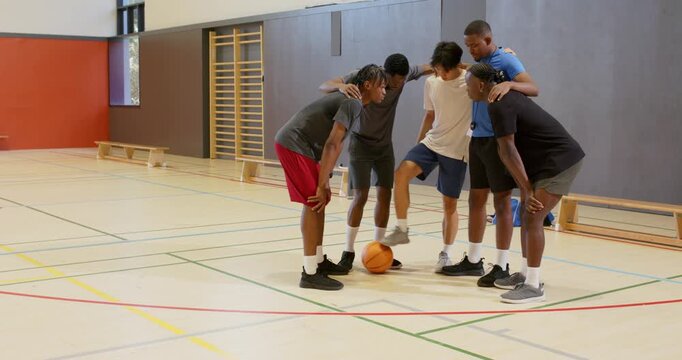 Coach passing basketball, diverse male team stacking hands, pumping arms for pep in school gym