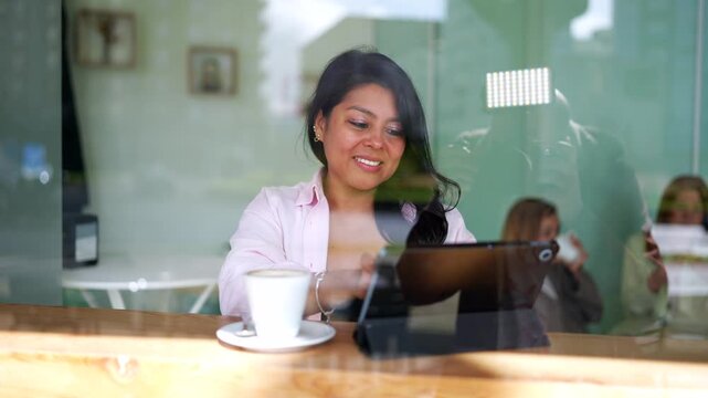 Young smiling asian businesswoman using a digital tablet while sitting at a wooden table in a coffee shop, enjoying her leisure time. View through the window with city life reflections