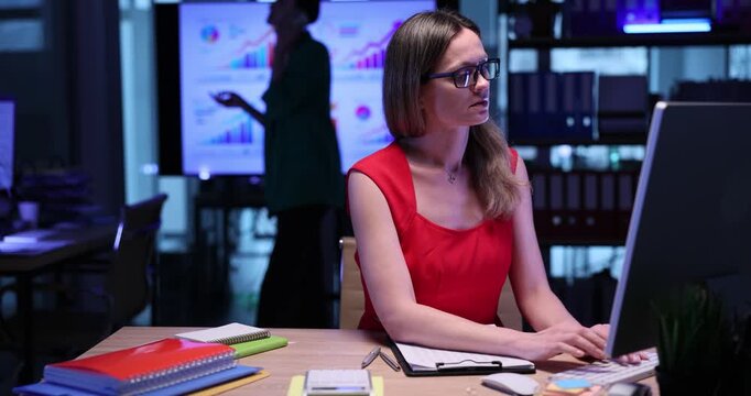 Woman manager with glasses types on computer keyboard and marks in checklist in semi-dark office. Blonde female employee checks data in workplace