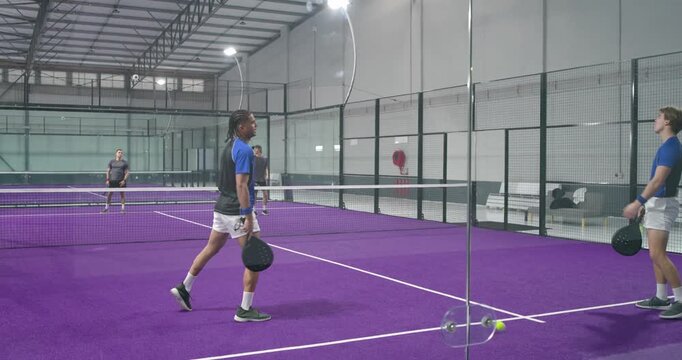 Diverse male friends exchanging padel volleys on purple court after far side lob shot for point