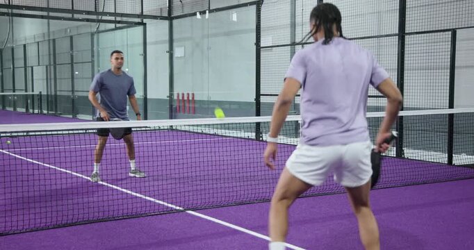 Male friends responding to lob over net on purple padel court while volleying yellow ball