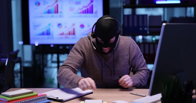 Fraud in mask finds opportunity to steal corporate financial data online in office. Man scammer with headset expresses joy and counts on calculator