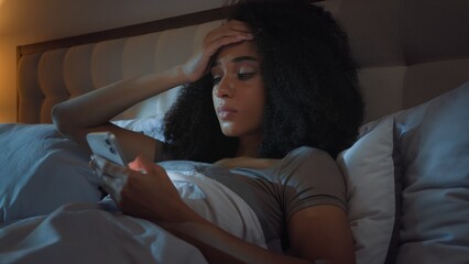Fototapeta premium Sad unwell African American girl scrolling mobile phone feeling headache at night in bed unhealthy stressed female woman using smartphone feel migraine frustrated with internet problem error cellphone