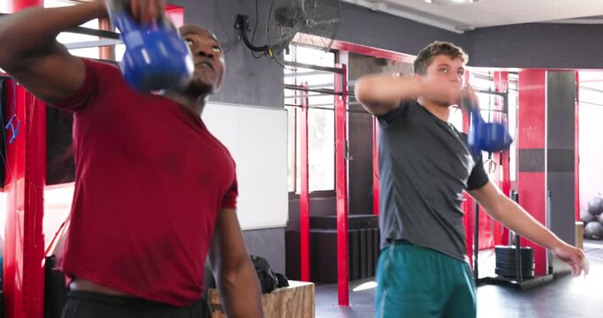Diverse male friends gripping kettlebells and hinging hips, swinging weights at crossfit gym