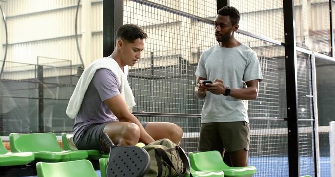 Diverse male friends exchanging juice, smartphone on court bench, viewing footage, shaking hands
