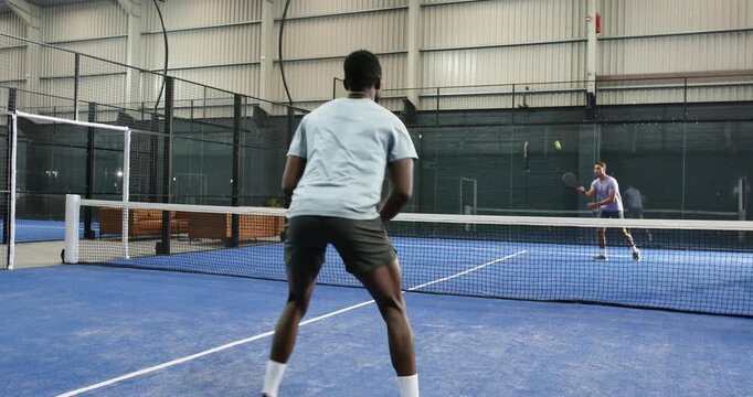 Diverse male athletes practicing padel rally after ball bounce on court inside