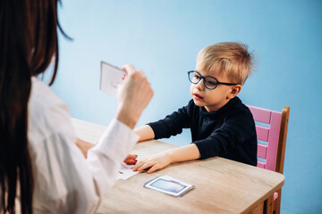 Preschool child with glasses engaged in learning activity with teacher at wooden table, colorful classroom background featuring blue wall and pink chair © standret