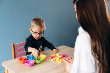 Obraz premium Preschool child with glasses engaged in colorful play activity with teacher at a wooden table, various bowls of materials arranged in front of them