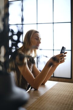 A woman sits at a wooden table looking at her smartphone. She is near a large window that lets in sunlight. The room has a simple design with minimal decorations and a relaxed atmosphere