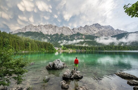 Young man standing on a rock on the shore, Eibsee lake in front of Zugspitze massif with Zugspitze, dramatic Mammaten clouds, Wetterstein range, near Grainau, Upper Bavaria, Bavaria, Germany