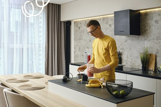 In a sleek kitchen, a man in a yellow shirt blends ingredients for a smoothie. He stands at a well-lit counter with fruits, a glass, and kitchen appliances around him, engaged in healthy cooking