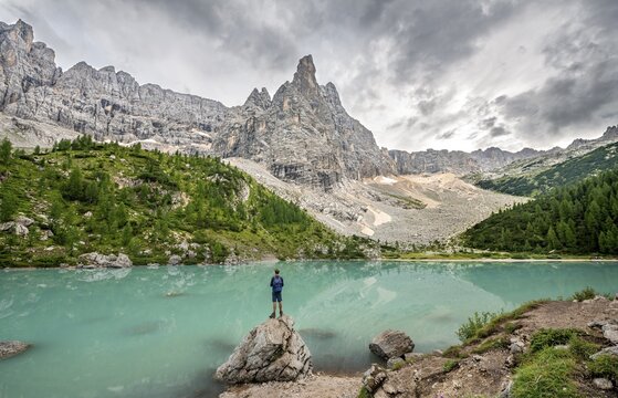 Young man standing on stone, hiker at the turquoise green Sorapis lake, Lago di Sorapis, mountain peak Dito di Dio, Dolomites, Belluno, Italy