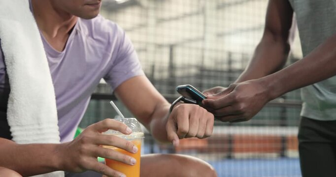 Diverse male duo receiving juice, syncing smartwatch for timing, shaking hands seat, copy space