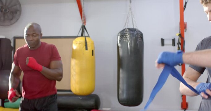 Diverse male partners wrapping hand wraps while shadowboxing by punching bag before workout at gym