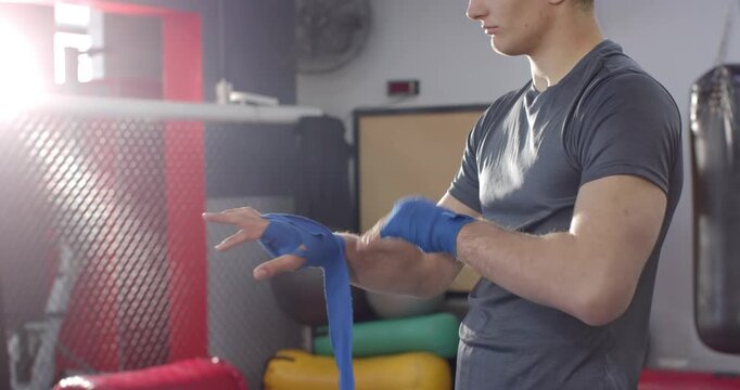 White male athlete gripping blue hand wrap and winding around wrists before boxing training in gym