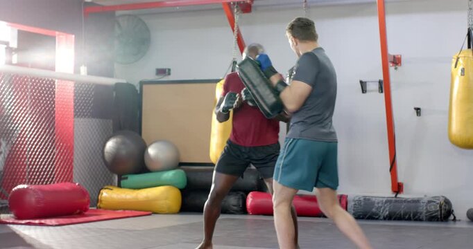 African American man at gym executing front kick and striking, training with coach raising mitts