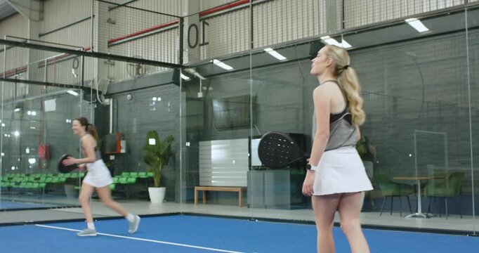 Baseline shot sending female rivals volleying on indoor padel court to sustain rally, chasing high l
