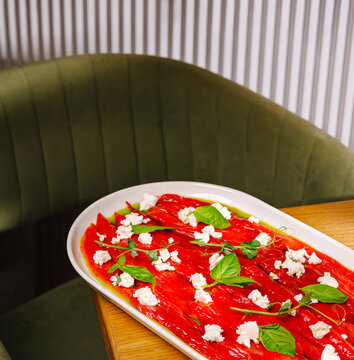 Delicious roasted red peppers carpaccio is served on white plate. Gourmet restaurant appetizer is decorated with crumbled feta cheese and fresh basil leaves.