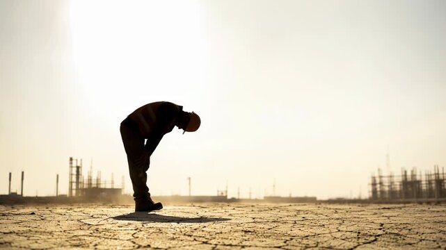 Silhouette of construction worker bending over cracked earth under extreme heat, showcasing harsh conditions and struggle of labor