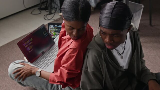 Young African American programmers sitting back-to-back on floor, writing code on laptops and discussing project. Footage for IT education, tech startups, and software development ads