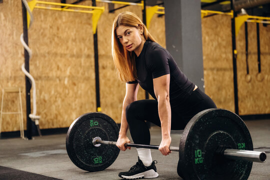 Female athlete with long blonde hair prepares to lift a barbell loaded with weights in a gym setting, with wooden walls and exercise equipment in the background