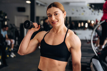 Fototapeta premium Female fitness enthusiast with muscular build holds dumbbell in gym, workout equipment and people exercising visible in the background