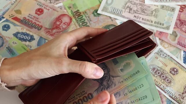 Vietnamese dong currency cash money close up. Person hands opening a brown leather wallet and pulling out green VND banknotes over a background scattered with different Vietnam bills, top view.