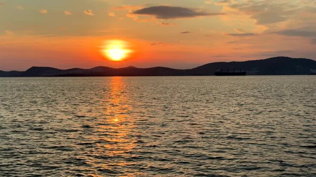 A calm, warm sunset over the Aegean Sea in Gulluk, Turkey. The sun hangs low in a hazy orange sky, casting a bright golden light path across the gently rippling water toward distant dark hills.