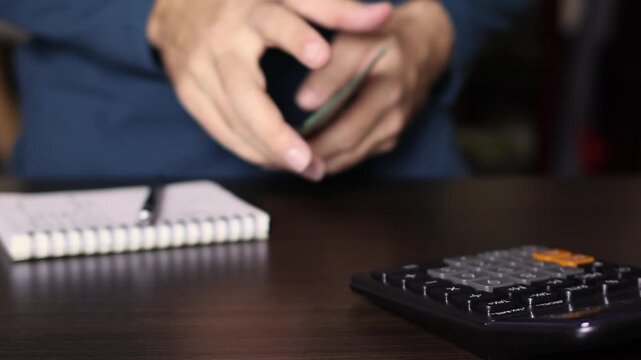 Close up of calculator on a wooden table with notebook and a businessman counting cash money bills in blurry background, selective focus. Financial accounting and budget planning concept.