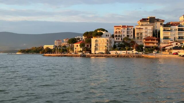 A beautiful evening view of Gulluk, Turkey, showing sunlit waterfront hotels and villas built on a hillside. The golden hour light reflects softly on the calm Aegean Sea near a quiet beach.
