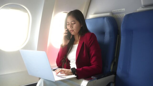 Businesswoman sitting by an airplane window, talking on her phone and using a laptop during a flight. Travel, remote work, business trip and focused multitasking concept.