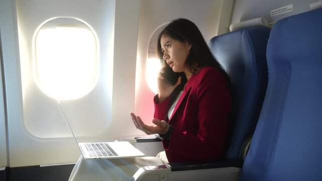 Businesswoman sitting by an airplane window, talking on her phone and using a laptop during a flight. Travel, remote work, business trip and focused multitasking concept.