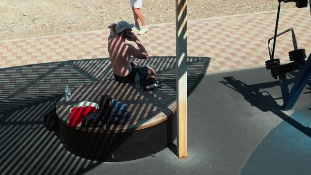Anonymous fit shirtless guy resting on round wooden bench under slatted shade after outdoor gym workout. Talking on mobile phone with strong striped shadow pattern on his back during summer day.