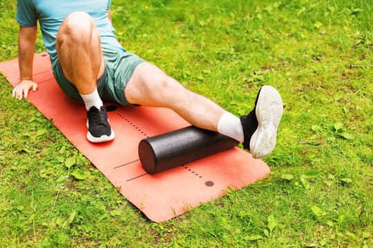 A man performs foam rolling on a mat for myofascial release and post-workout recovery, using a roller for leg muscle self-massage outdoors in a park.