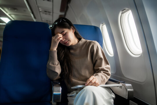 Tired young woman sitting by an airplane window, rubbing her forehead during a flight. Travel stress, fatigue, jet lag and discomfort inside a modern passenger cabin.